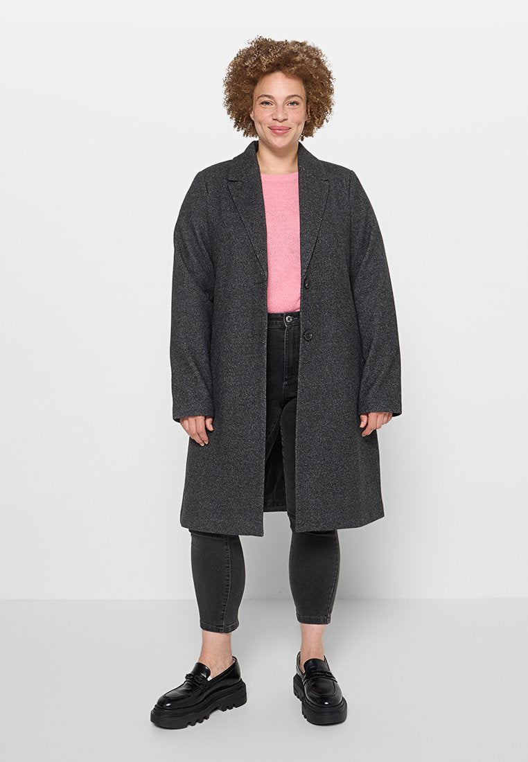Langer grauer Mantel mit zwei vorderen Knöpfen, über einem rosa Oberteil und dunklen Jeans getragen. Verfügt über einen lässigen Schnitt und strukturierter Stoff. Schwarze Schuhe runden den Look ab.