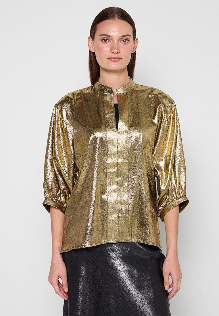 Frau mit glattem braunem Haar, die eine glänzende goldene Bluse mit langen Ärmeln und Schlitz am Ausschnitt sowie einen schwarzen strukturierten Rock trägt.