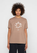 Frau mit lockigem Haar, die ein braunes T-Shirt mit weißem kreisförmigem Text „LISA/SAINT“ und „LONDON“ trägt, kombiniert mit schwarzen Hosen, steht vor weißem Hintergrund.