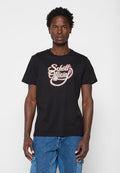 Mann mit kurzen lockigen Haaren, der ein schwarzes T-Shirt mit dem Text "Schott Official" und blaue Jeans trägt, steht vor einem schlichten weißen Hintergrund.