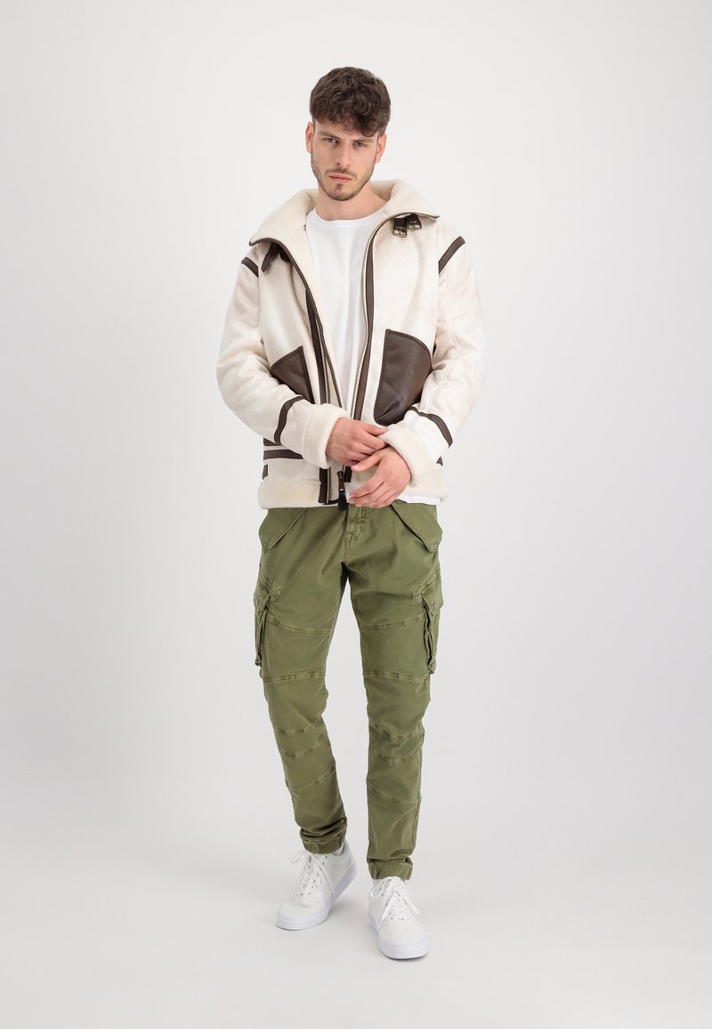 Cremfarbener und brauner Reißverschlussjacke mit einem pelzähnlichen Textur, die große Taschen und eine Kapuze hat. Kombiniert mit olivfarbenen Cargo-Hosen und weißen Turnschuhen.
