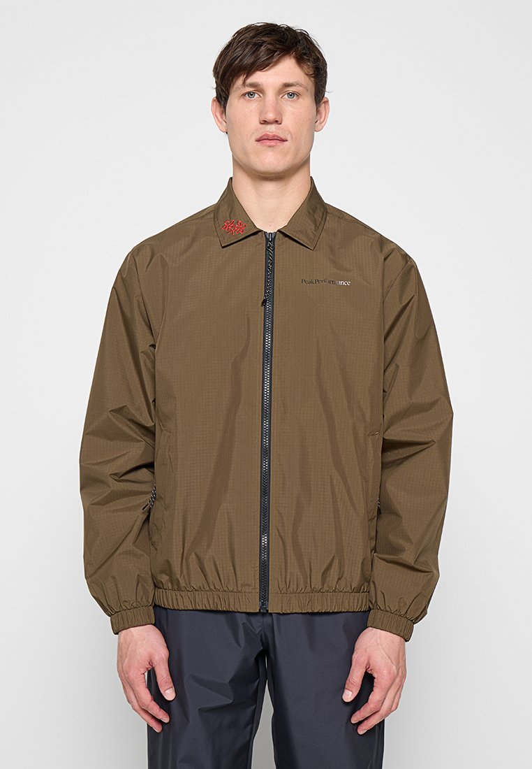 Junger Mann trägt eine olivgrüne Jacke mit Reißverschluss und elastischen Bündchen, steht vor einem einfarbigen hellen Hintergrund, blickt geradeaus mit neutralem Gesichtsausdruck.