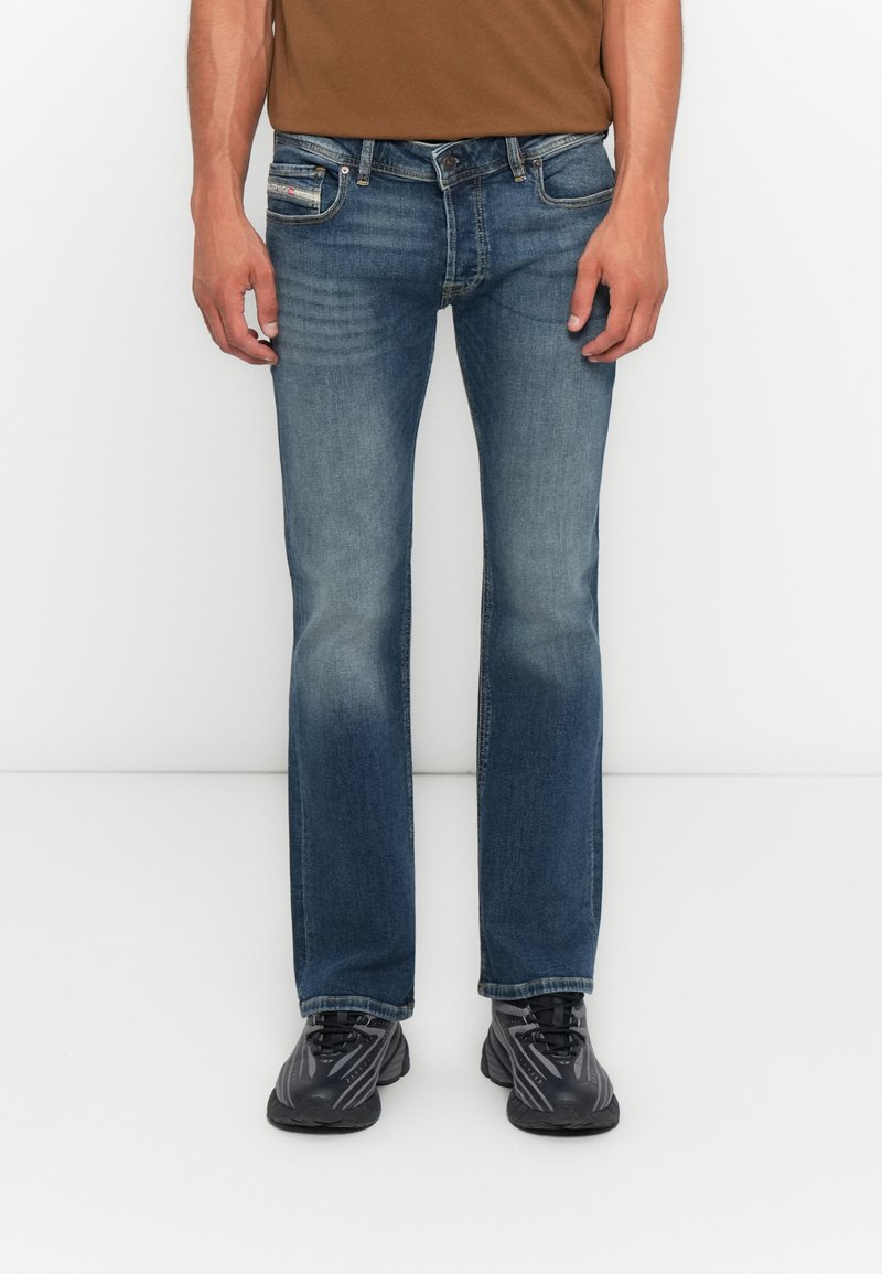 Mann in ausgebleichten blauen Straight-Leg-Jeans und schwarzen Sneakers, der vor einem schlichten weißen Hintergrund steht.