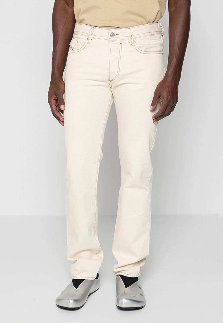 Beige Baumwolljeans mit geradem Bein, kontrastierenden orangen Nähten und Fünf-Taschen-Design, getragen mit silbernen Slip-On-Schuhen.