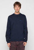 Mann trägt einen dunkelblauen Langarm-Sweatshirt mit kleinem blauem Text auf der Brust und dunkelroten gewaschenen Jeans vor einem einfarbigen Hintergrund.