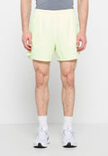 Mann trägt blassgelbe Sportshorts, weiße Socken und weiße Laufschuhe und steht vor einem schlichten hellen Hintergrund.