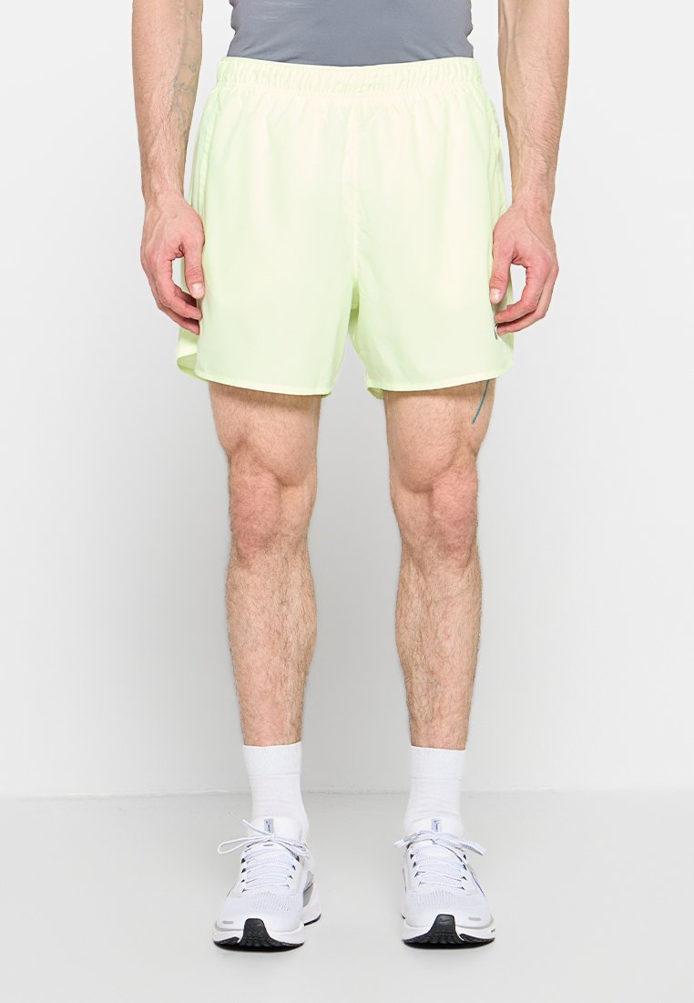 Mann trägt blassgelbe Sportshorts, weiße Socken und weiße Laufschuhe und steht vor einem schlichten hellen Hintergrund.