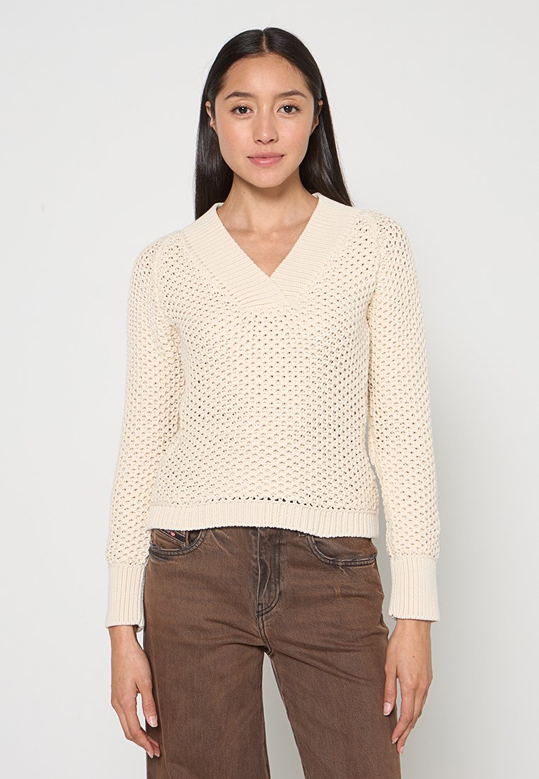 Cremefarbener, gestrickter Pullover mit V-Ausschnitt und langen Ärmeln. Verfügt über ein strukturiertes Muster sowie gerippte Bündchen und Saum. Wird mit braunen Hosen kombiniert.