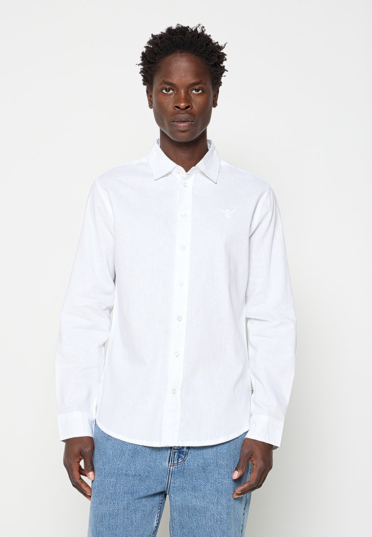 Mann mit kurzen lockigen Haaren, der ein weißes langärmeliges Hemd und blaue Jeans trägt, steht vor einem schlichten weißen Hintergrund.
