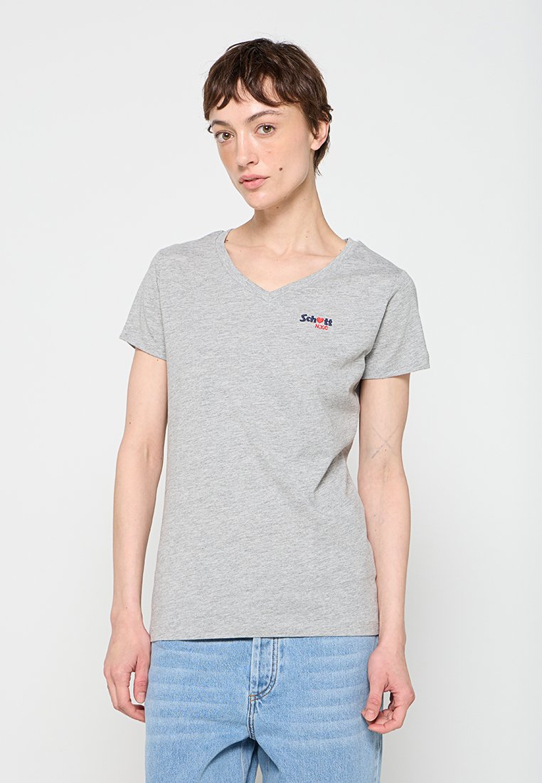 Junge Person mit kurzen braunen Haaren, die ein hellgraues V-Ausschnitt-T-Shirt und hellblaue Jeans trägt, steht vor einem schlichten weißen Hintergrund.