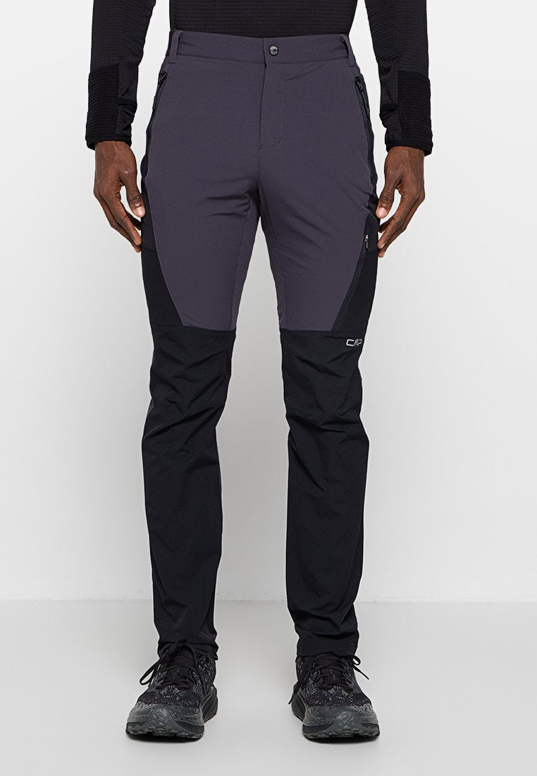 Mann trägt zweifarbige schwarze und dunkelgraue Outdoorhose mit Reißverschlusstaschen an den Seiten und schwarzen Sportschuhen, steht vor einem einfarbigen Hintergrund.