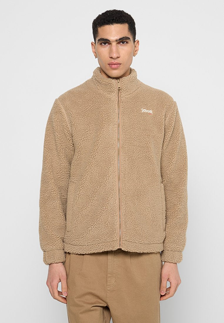 Mann in beiger Fleecejacke mit Reißverschluss und passenden Hosen, der vor einem schlichten hellgrauen Hintergrund steht.