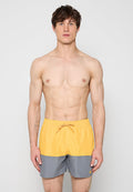 Oberkörperfreier Mann mit kurzen braunen Haaren, der gelbe und graue Badeshorts trägt und vor einem schlichten hellen Hintergrund steht.