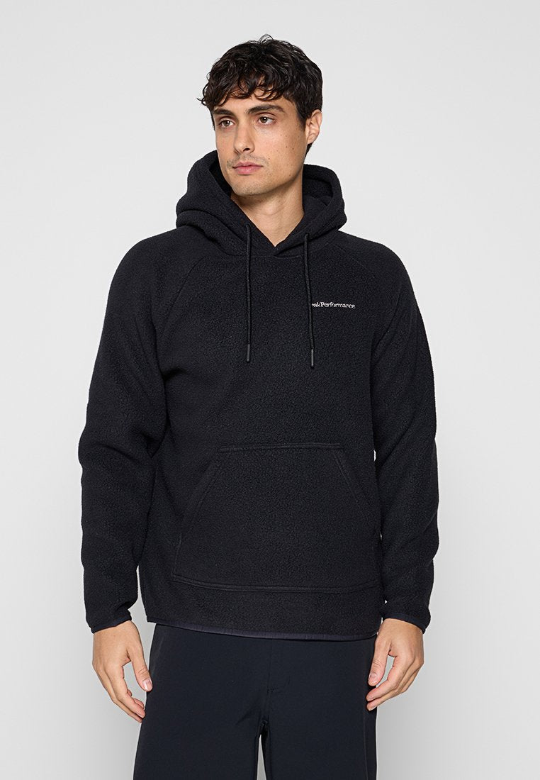 Mann trägt einen schwarzen Fleece-Kapuzenpullover mit Fronttasche und kleinem weißen Logo auf der Brust, steht vor grauem Hintergrund.
