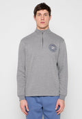 Mann trägt einen hellgrauen Pullover mit Viertel-Reißverschluss und einem runden Logo auf der linken Brust sowie blaue Hosen, steht vor einem schlichten Hintergrund.