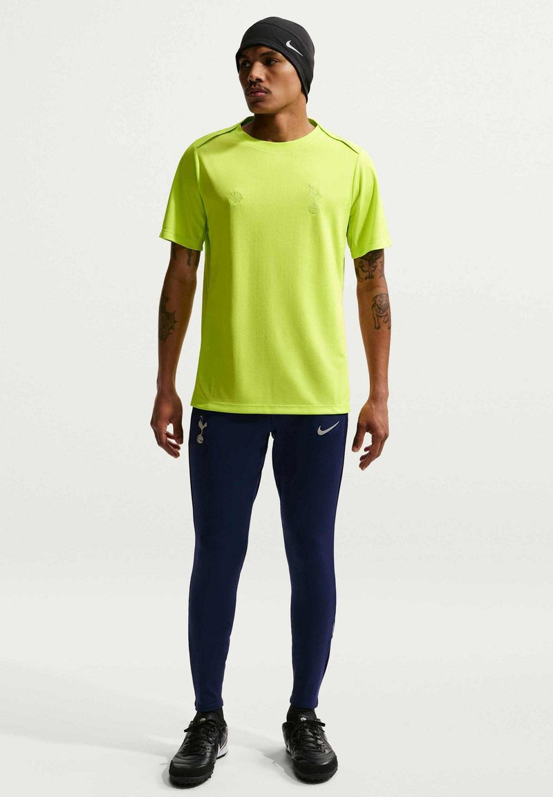 Mann trägt neon-gelbes kurzärmeliges Hemd, dunkelblaue Sporthose, schwarze Turnschuhe und eine schwarze Mütze, steht vor weißem Hintergrund.