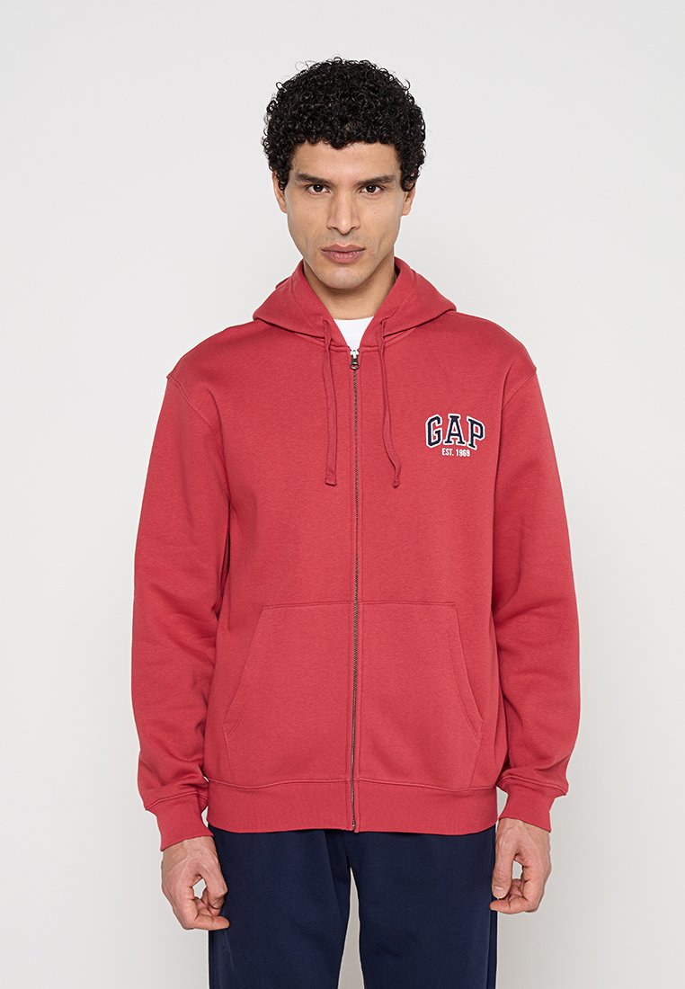 Mann trägt einen roten Kapuzenpullover mit Reißverschluss und der Aufschrift "GAP EST. 1969" auf der Brust, steht vor einem schlichten, hellen Hintergrund.