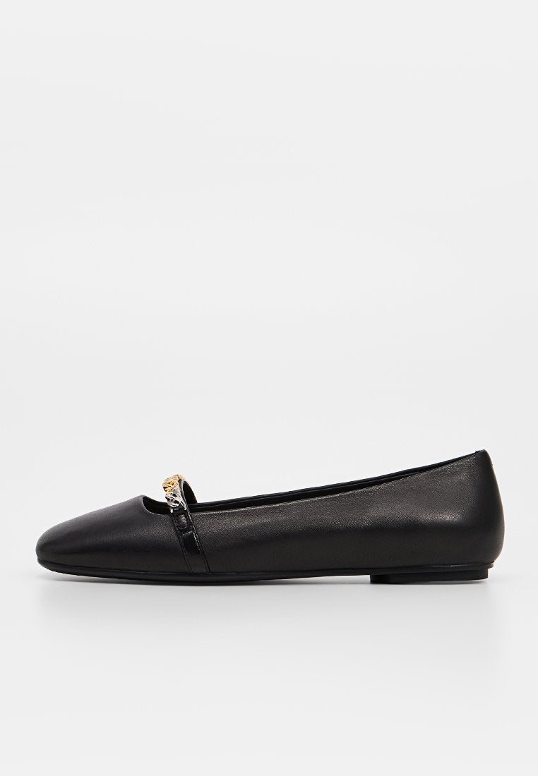 Schwarzer flacher Schuh mit spitzer Spitze und kleinem goldenen Ketten-Detail oben, fotografiert vor einfachem weißem Hintergrund.
