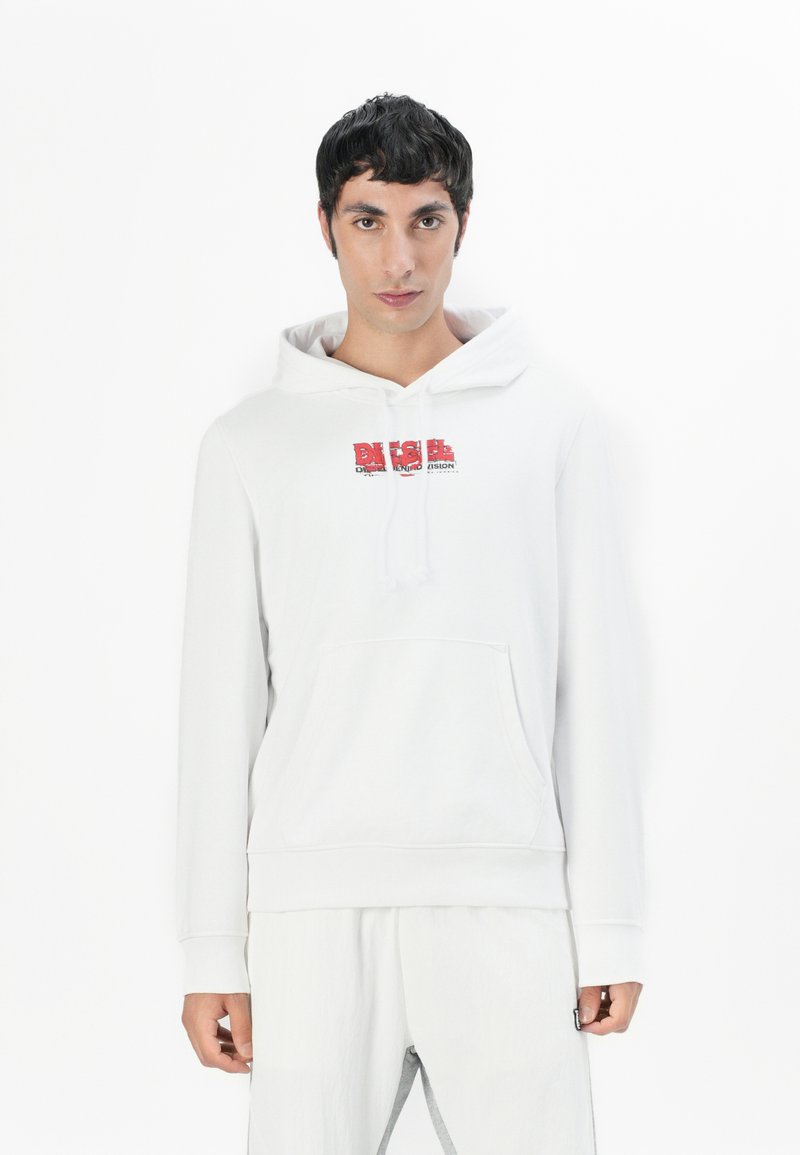 Mann in weißem Hoodie mit rotem Diesel-Logo, weißen Hosen, steht vor einem schlichten weißen Hintergrund und blickt nach vorne.