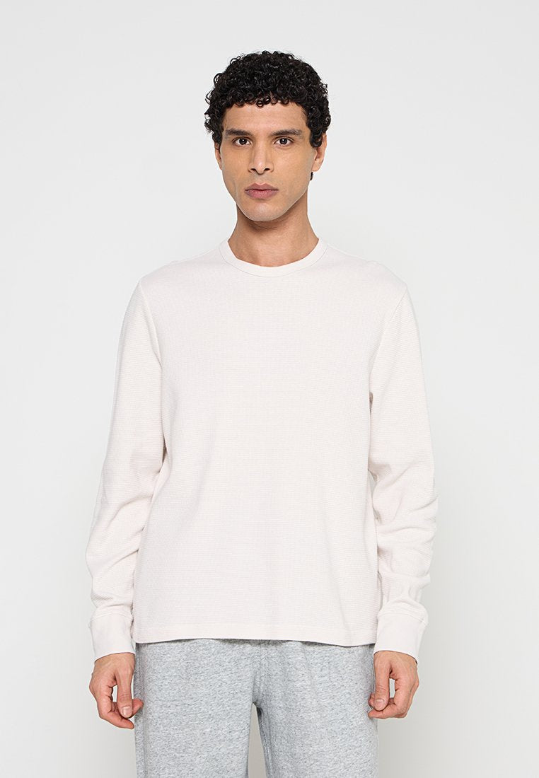 Mann mit lockigem dunklem Haar, der ein einfarbiges weißes Langarmshirt und hellgraue Hosen trägt, vor einem weißen Hintergrund steht.