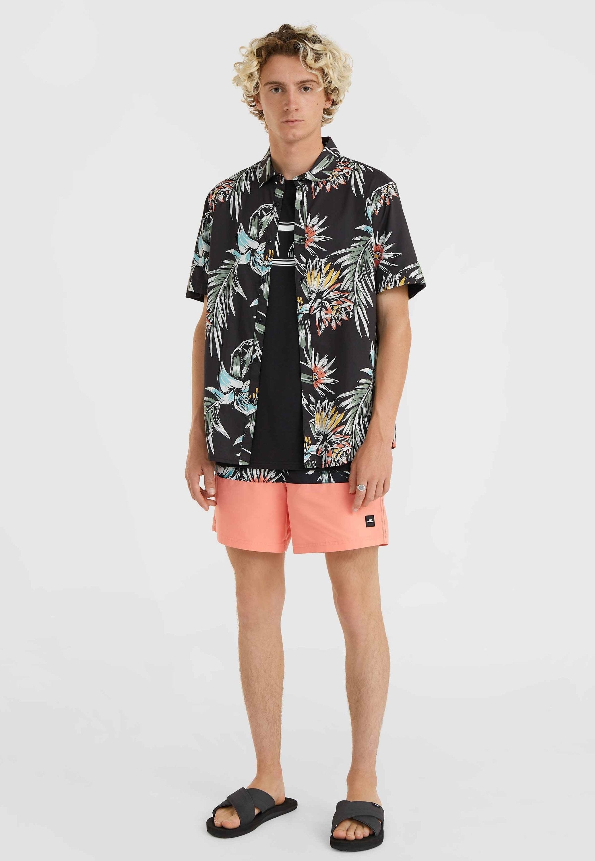 Kurzärmliges schwarzes Blumenhemd mit bunten Blattformen, kombiniert mit korallenfarbenen Badeshorts und schwarzen Sandalen, zeigt legere Sommermode.