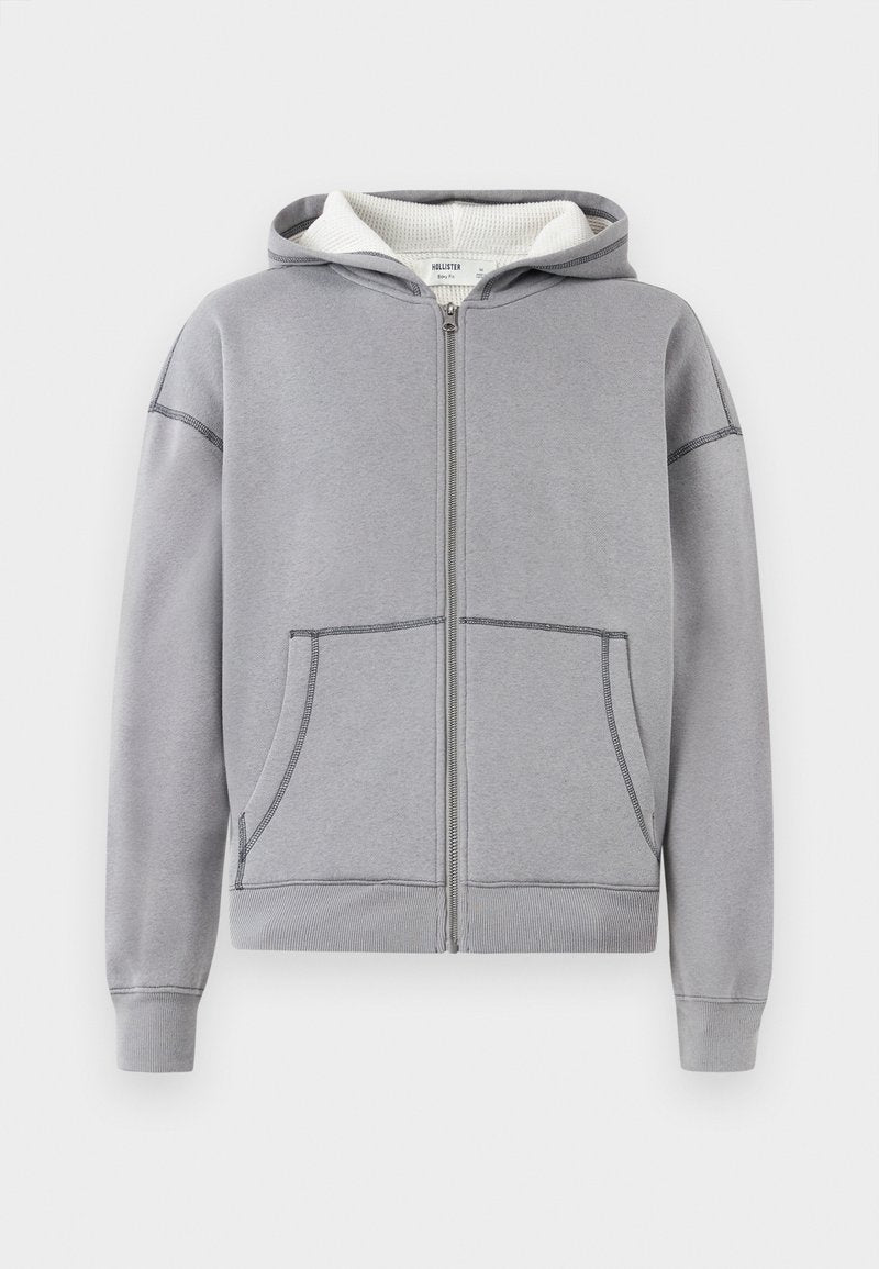 Grauer Hoodie mit Reißverschluss, Fronttaschen, gerippten Bündchen und Saum, strukturiertem weißen Innenfutter und kontrastierenden Detailnähten.