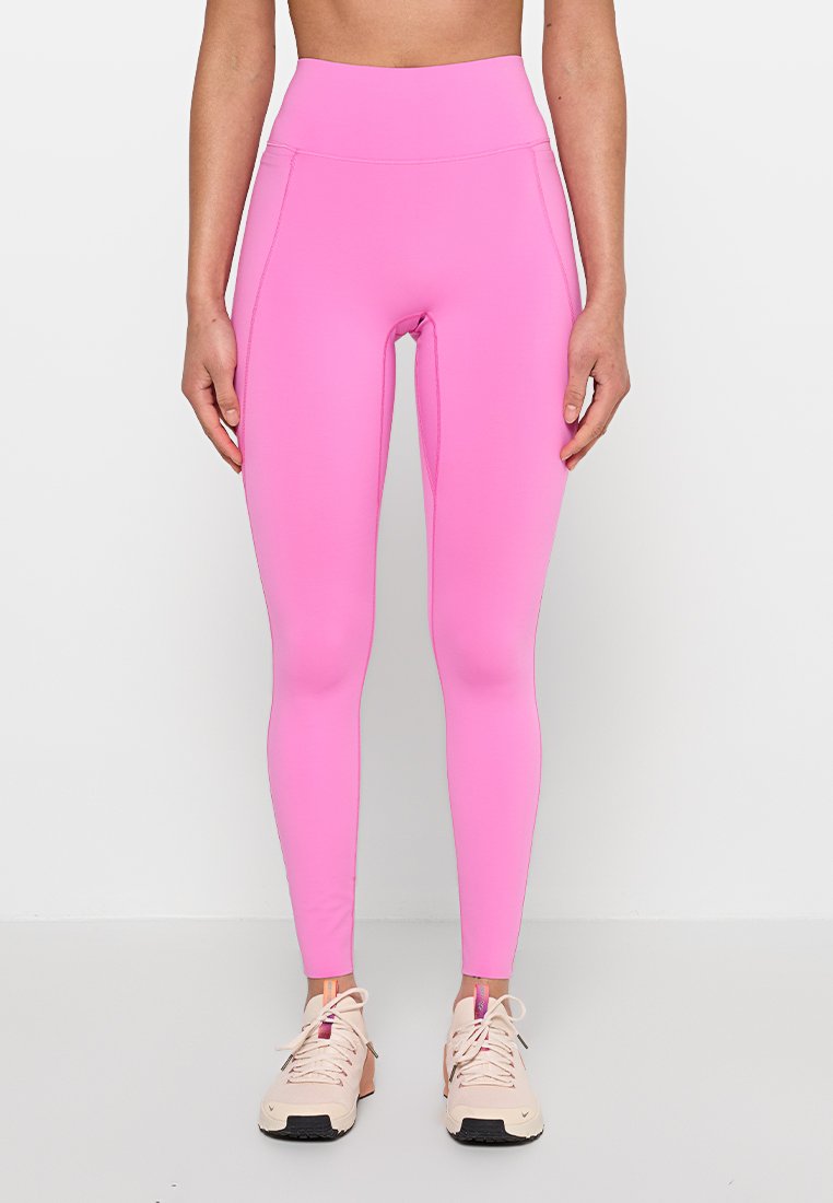 Frau trägt leuchtend pinke High-Waist-Leggings und hellrosa Sportchuhe, steht vor einem schlichten weißen Hintergrund.