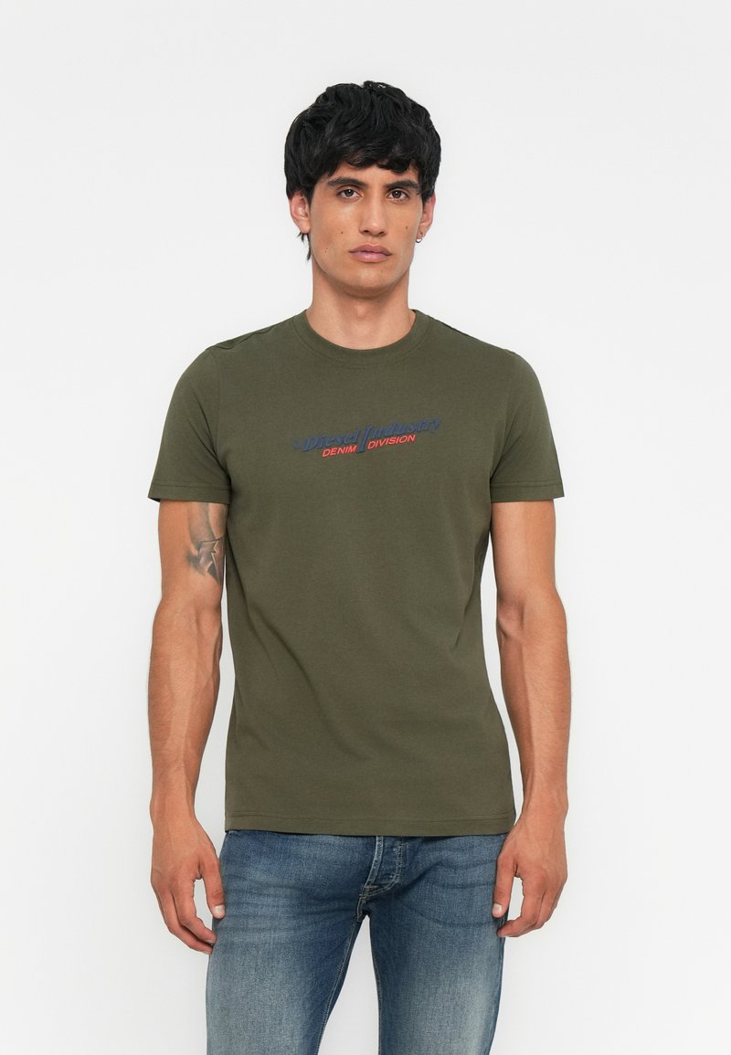 Olivegrünes Baumwoll-T-Shirt mit kurzen Ärmeln, Rundhalsausschnitt und bedrucktem Logo in Blau und Rot. Modell steht in eng anliegenden blauen Jeans.