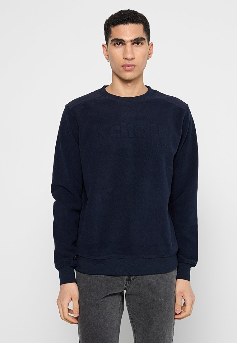Junger Mann in einem dunkelblauen Sweatshirt mit dezentem "Schott NYC"-Logo, dunklen grauen Jeans, stehend vor einem schlichten hellen Hintergrund.