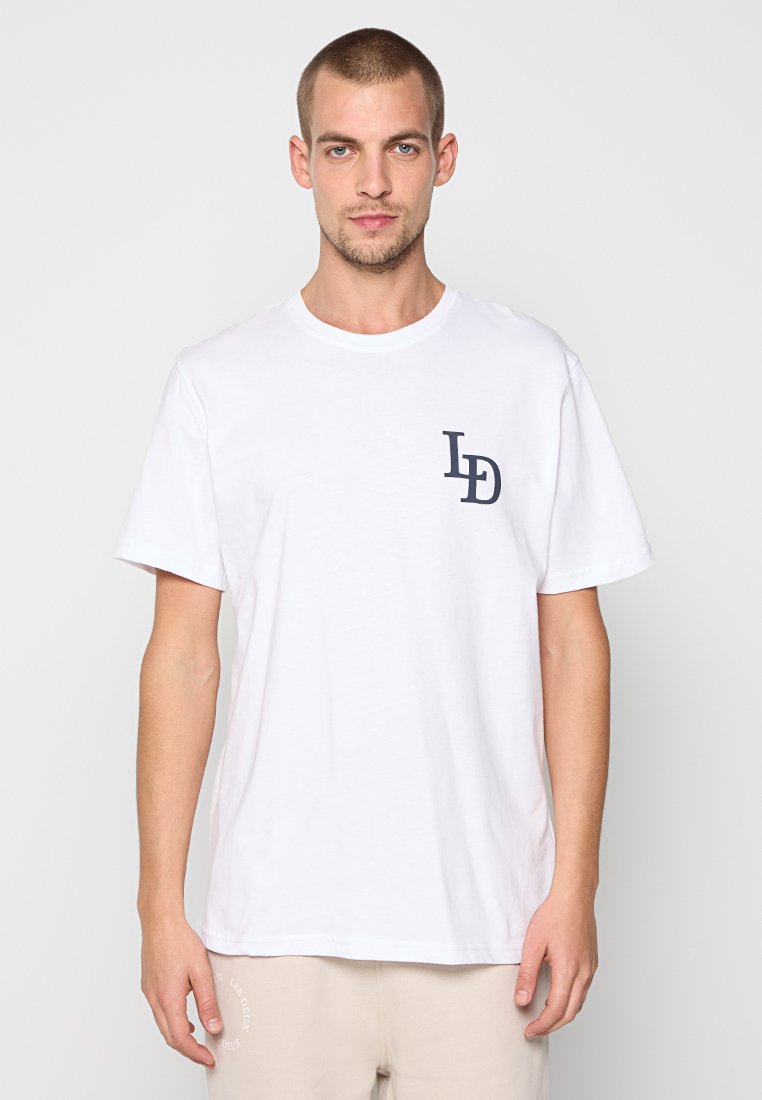 Mann mit kurzen Haaren, der ein weißes T-Shirt mit den Buchstaben "LD" auf der Brust und beigefarbenen Hosen trägt, steht vor einem schlichten, hellen Hintergrund.