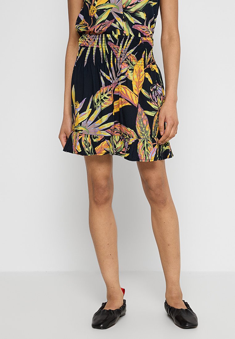 Blumenmusterkleid mit dunklem Hintergrund, geraffter Taille, ausgestelltem Saum und bunten Blattmustern. Begleitet von schwarzen flachen Schuhen.