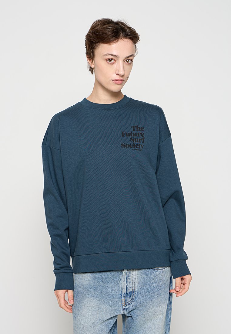 Person mit kurzen braunen Haaren, die einen blauen Pullover mit schwarzem Schriftzug und hellblaue Jeans trägt, steht vor einem einfarbigen hellen Hintergrund.