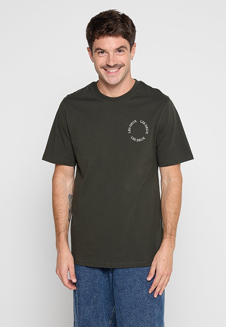 Dunkelgrünes Baumwoll-T-Shirt mit Rundhalsausschnitt und kurzen Ärmeln. Mit einem weißen, runden Logo-Druck im linken Brustbereich.