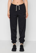 Person trägt dunkelgraue Sweatpants mit weißem Kordelzug und schwarzen Sneakers, steht vor einem schlichten hellen Hintergrund.
