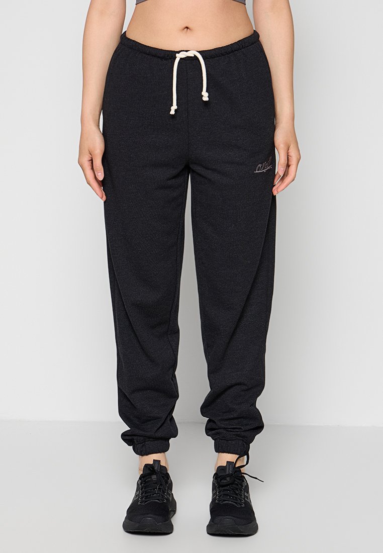 Person trägt dunkelgraue Sweatpants mit weißem Kordelzug und schwarzen Sneakers, steht vor einem schlichten hellen Hintergrund.