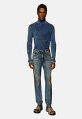 Mann in einem taillierten blauen gerippten Rollkragenpullover, ausgebleichten blauen Jeans und schmutzigen schwarzen Stiefeln mit metallischen Zehenschutzkappen, vor einem weißen Hintergrund stehend.