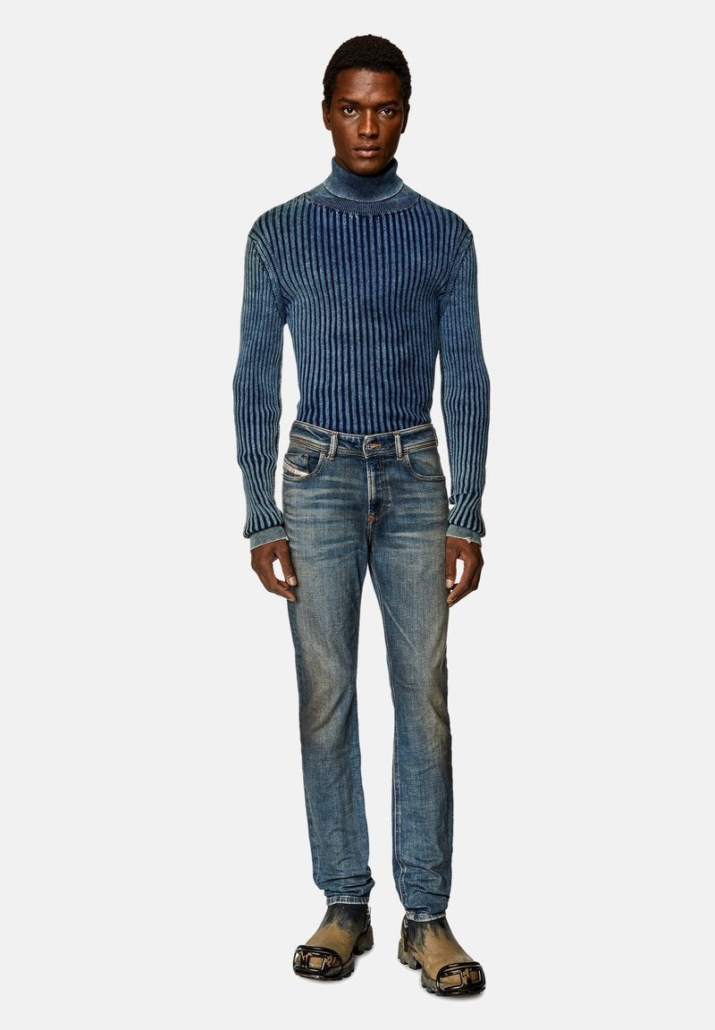 Mann in einem taillierten blauen gerippten Rollkragenpullover, ausgebleichten blauen Jeans und schmutzigen schwarzen Stiefeln mit metallischen Zehenschutzkappen, vor einem weißen Hintergrund stehend.