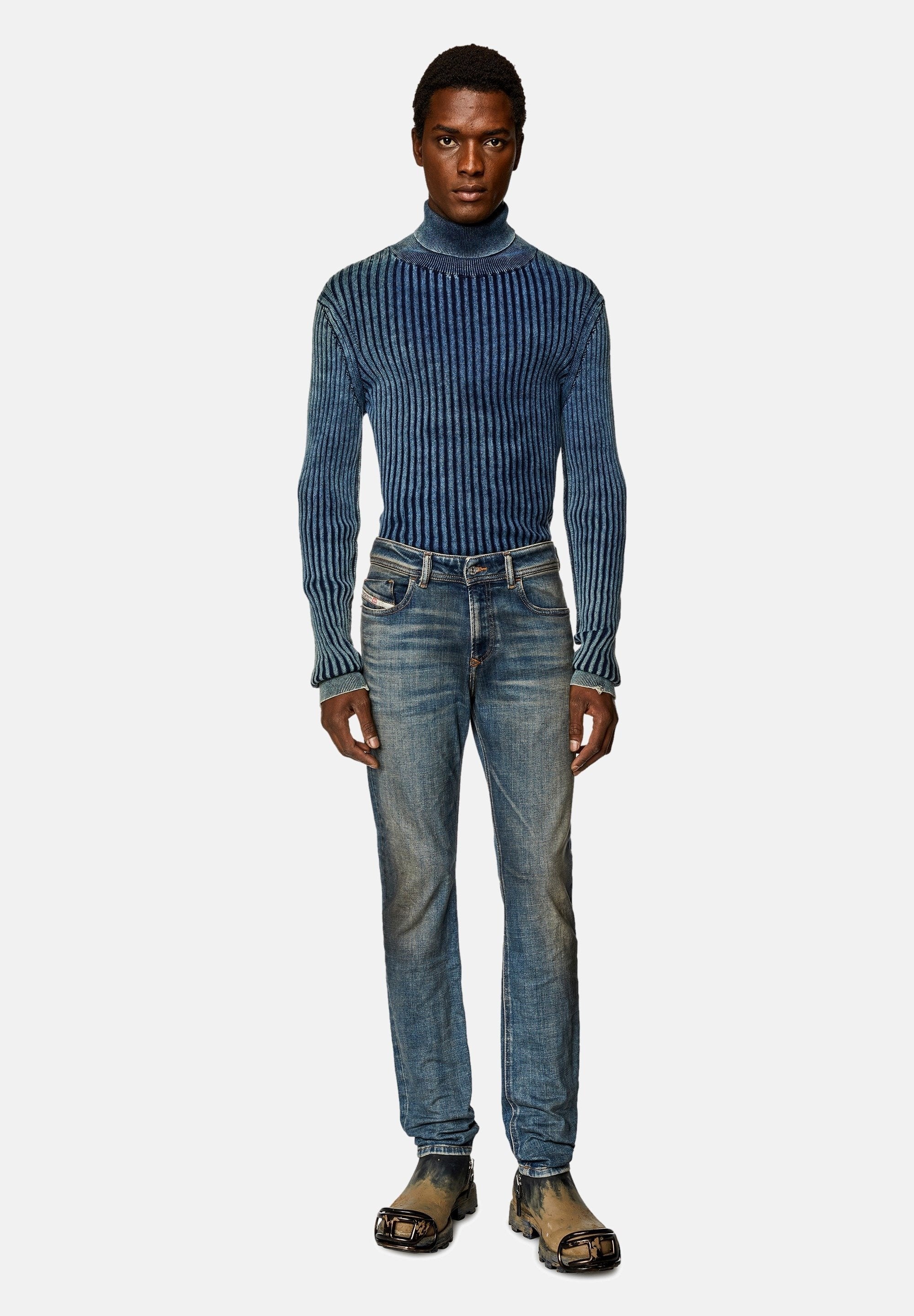 Mann in einem taillierten blauen gerippten Rollkragenpullover, ausgebleichten blauen Jeans und schmutzigen schwarzen Stiefeln mit metallischen Zehenschutzkappen, vor einem weißen Hintergrund stehend.