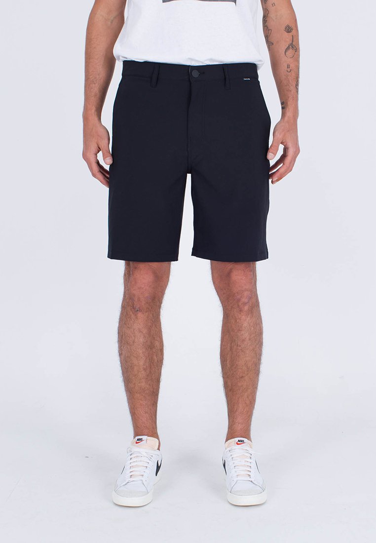 Mann in schwarzen knielangen Shorts, weißen Nike-Sneakern und einem weißen T-Shirt, der vor einem schlichten hellen Hintergrund steht.
