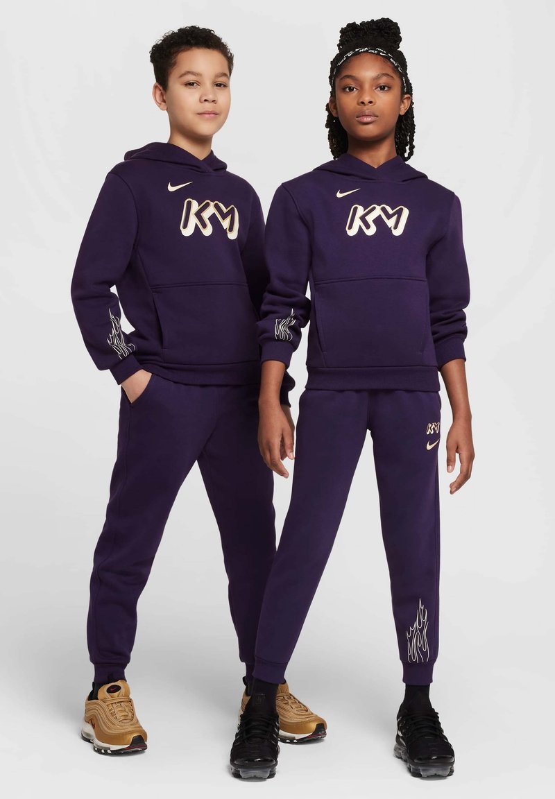 Lila Kapuzenpullover und -hosen mit weißem "KM"-Logo. Die Hosen verfügen über ein Flammendesign an den Säumen. Die Schuhe des Modells sind schwarz-goldene Sneaker.