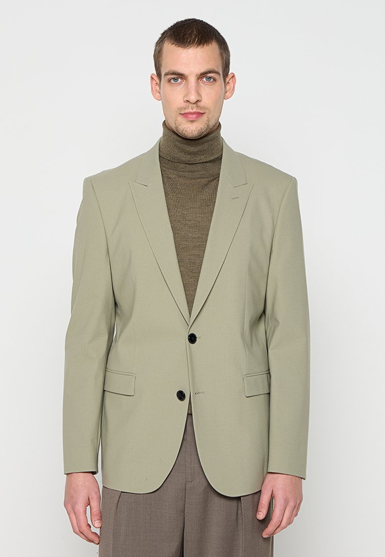 Hellgrüner Blazer mit strukturiertem Design, zwei schwarzen Knöpfen und Vordertaschen, getragen über einem dunkelbraunen Rollkragenpullover.