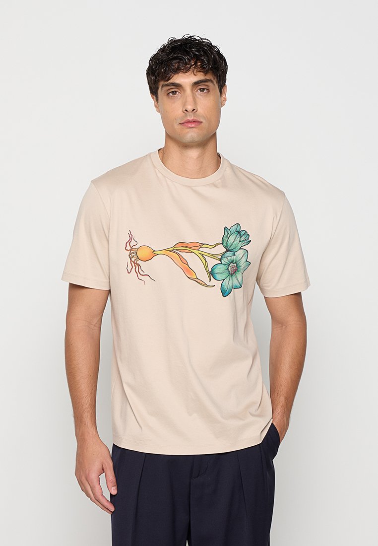 Junger Mann mit kurzem dunklem Haar, der ein beiges T-Shirt mit einem bunten botanischen Design mit blauen Blumen und einer orangefarbenen Knolle trägt.