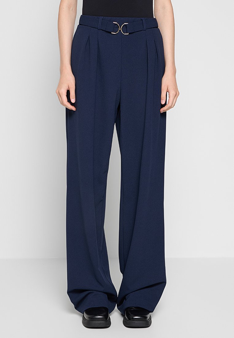 Marineblaue weite Hosen verfügen über einen vorderen Faltenwurf, einen Gürtel mit einer goldenen kreisförmigen Schnalle und eine glatte Stofftextur. Kombiniert mit schwarzen Schuhen.