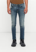 Mann in schmal geschnittenen blauen Jeans und schwarzen Sportschuhen, der gegen einen weißen Hintergrund steht, mit entspannten Händen an den Seiten.