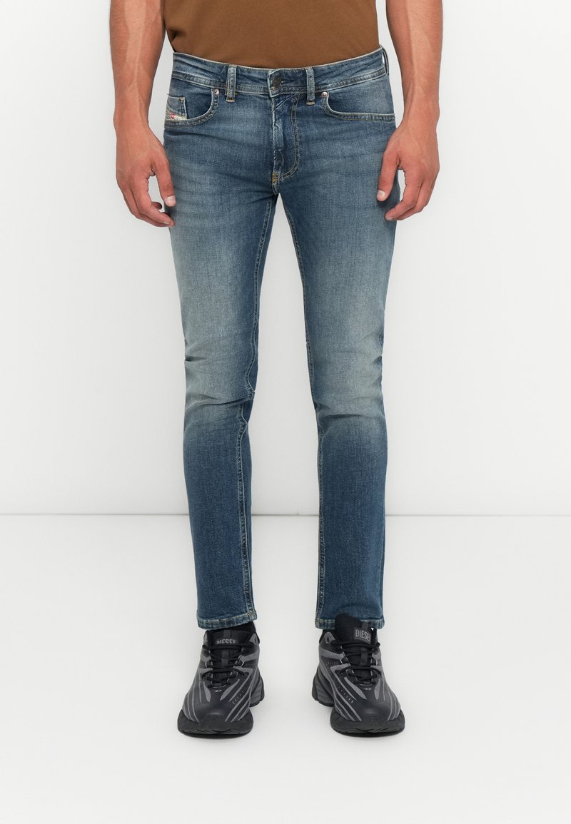 Mann in schmal geschnittenen blauen Jeans und schwarzen Sportschuhen, der gegen einen weißen Hintergrund steht, mit entspannten Händen an den Seiten.