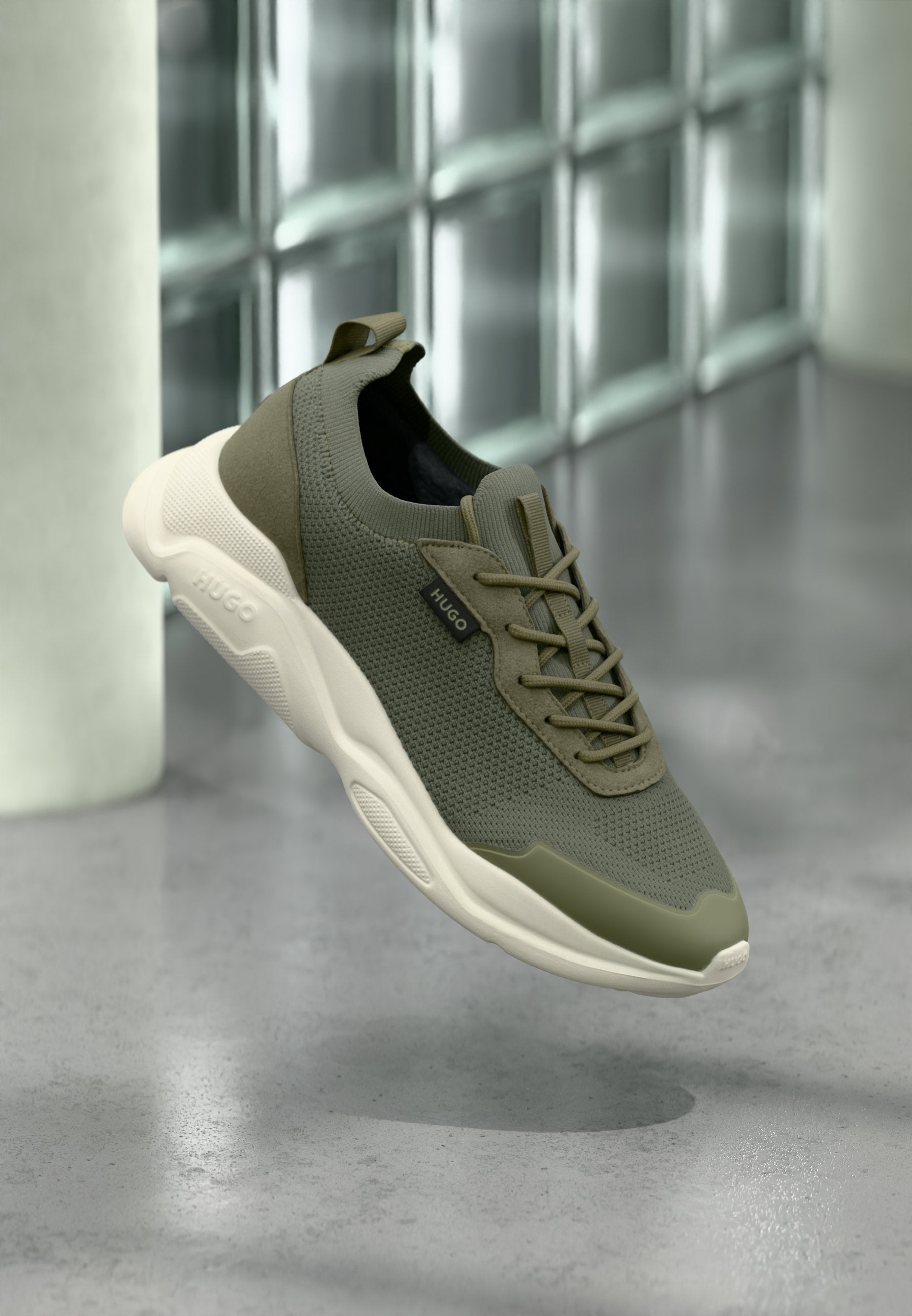 Olivgrüner Strick-Sneaker mit weißer, klobiger Sohle und "HUGO"-Label, der schwebend über einem grauen, polierten Boden nahe Glasbausteinen gezeigt wird.