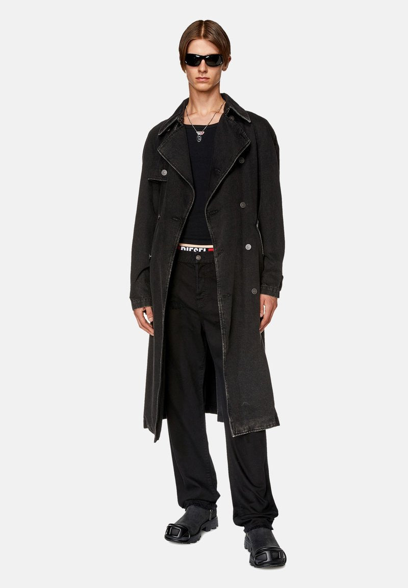 Schwarzer Denim-Trenchcoat mit großen Revers und doppelter Knopfleiste. Getragen über einem schwarzen Oberteil und Hose, ergänzt durch klobige Schuhe.