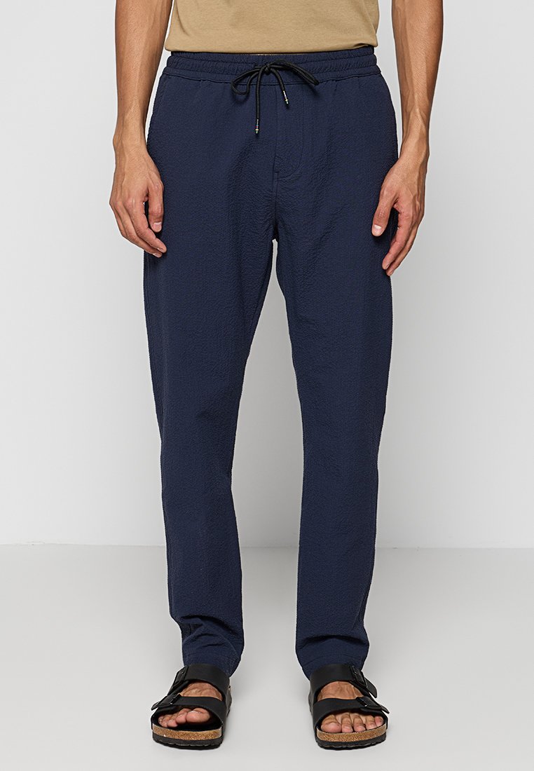 Marineblaue, leichte, strukturierte Hosen mit elastischem Bund und verstellbarem Kordelzug, kombiniert mit schwarzen Sandalen mit Schnalle.