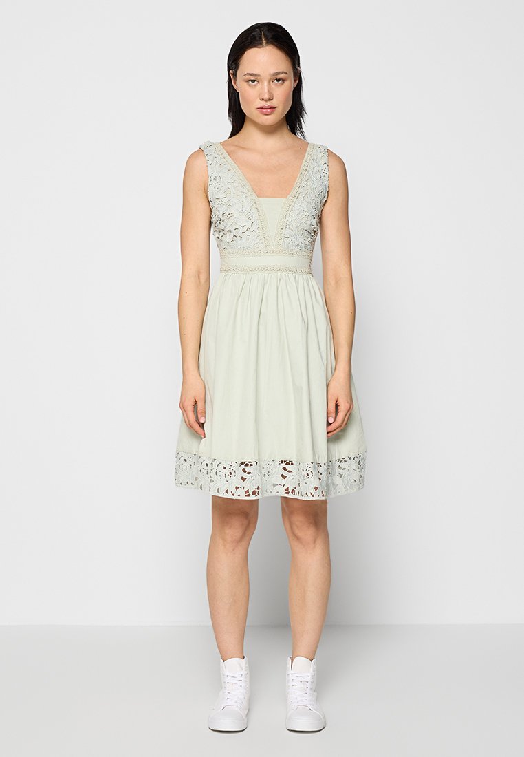 Hellgrünes Kleid mit einem taillierten Oberteil und schwingendem Rock, verziert mit Spitze am Ausschnitt und dem Saum. Kombiniert mit weißen Sneakers.