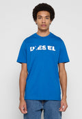 Junger Mann mit lockigem Haar, der ein leuchtend blaues Diesel-T-Shirt und blaue Jeans trägt, vor einem schlichten hellgrauen Hintergrund steht.